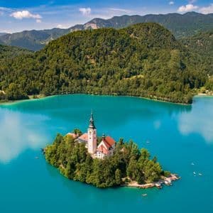 Una vista aérea de una iglesia en una pequeña isla cubierta de árboles en medio de un lago turquesa, con montañas boscosas en el fondo.