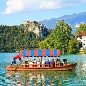 Un voyage de groupe WeRoad à bord d'un bateau en bois, naviguant sur un lac turquoise, avec un château niché sur une falaise boisée et des montagnes en arrière-plan.