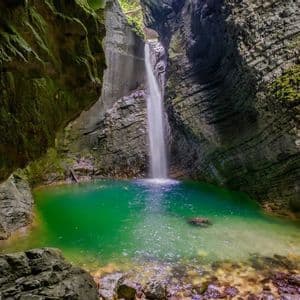 Une cascade se jette dans un bassin d'eau turquoise éclatante, entourée de parois rocheuses abruptes et couvertes de mousse dans une gorge.