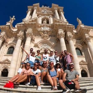 Un gruppo WeRoad in viaggio posa per una foto sui gradini di pietra davanti a un sontuoso edificio barocco sotto un cielo azzurro.