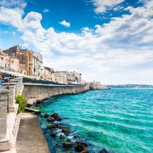 De vieux bâtiments d'une ville côtière bordent une digue de pierre le long d'une eau turquoise, sous un ciel bleu parsemé de nuages blancs.