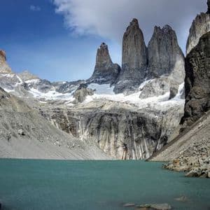 A turquoise glacial lake sits at the base of three jagged, towering granite peaks covered in snow under a blue sky with clouds.
