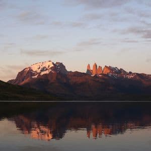 Jagged, snow-capped mountains are illuminated by sunrise, with their reflection visible in the still lake below.