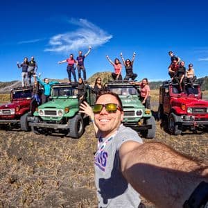 Un homme prend un selfie avec un groupe WeRoad posant sur et autour de jeeps tout-terrain colorées dans un vaste paysage aride sous un ciel bleu.