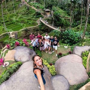 A woman takes a selfie with a WeRoad group trip, posing together on a hill overlooking lush green rice terraces and palm trees.