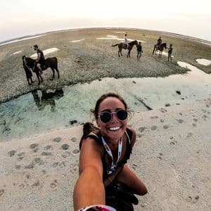 A smiling woman in sunglasses takes a selfie on a sandy beach while her WeRoad group trip rides horses in the background.