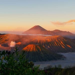 Une série de cratères et de montagnes volcaniques illuminés par la lumière chaude du lever du soleil, avec un cratère émettant un panache de fumée.