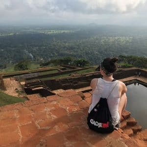 Una persona con una mochila WeRoad se sienta sobre ruinas de ladrillo con vistas a un vasto valle boscoso desde un mirador alto.