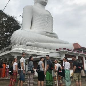 Un gruppo WeRoad in viaggio, a piedi nudi, guarda la fotocamera di fronte a una gigantesca statua bianca del Buddha seduto.