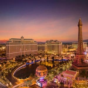 An aerial view of an illuminated cityscape featuring a replica Eiffel Tower and large hotels under a colorful sunset sky.
