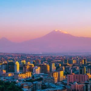 Un paysage urbain dense, avec des bâtiments éclairés par le coucher de soleil et une grande montagne à double sommet en arrière-plan sous un ciel coloré.