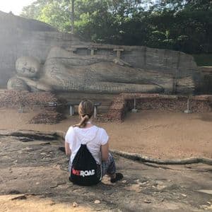 A person with a WeRoad backpack sits on a rock looking at a large reclining Buddha statue carved into a cliff face.