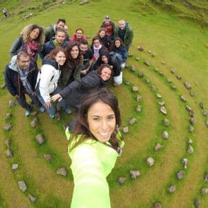 Une femme en veste fluo prend un selfie avec un voyage de groupe WeRoad à l'intérieur d'une spirale de pierres sur une colline verte.
