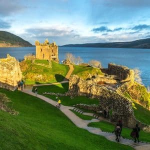 Des gens marchent sur des sentiers à travers les ruines d'un château en pierre sur une colline herbeuse surplombant un grand lac et des montagnes lointaines.