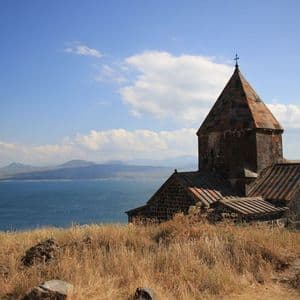 Une vieille église en pierre au toit de tuiles se dresse sur une colline herbeuse, dominant un grand lac avec des montagnes en arrière-plan.