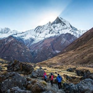 Eine WeRoad Gruppenreise von drei Personen, die bei Sonnenaufgang mit Wanderstöcken auf einem felsigen Pfad zu einem imposanten schneebedeckten Berg wandern.