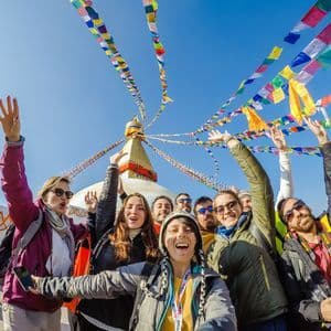 Un viaje en grupo de WeRoad se toma una selfie sonriente frente a una gran estupa bajo coloridas banderas de oración y un cielo azul claro.