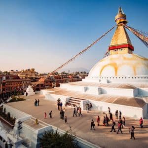 An elevated view of a large, white Buddhist stupa with a golden spire and prayer flags, as people walk on the terrace below.