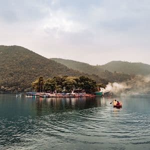 Una piccola barca con due persone viene remata su un lago verso un'isola alberata, con verdi colline sullo sfondo.