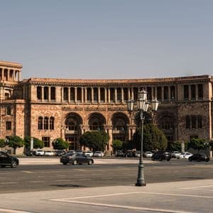 Un bâtiment monumental en pierre avec une tour d'horloge et des colonnades arquées se dresse sur une vaste place de ville avec des voitures.