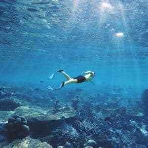 Eine Person mit Flossen schnorchelt über einem riesigen Korallenriff in klarem blauem Wasser, wobei Sonnenstrahlen durch die Oberfläche brechen.