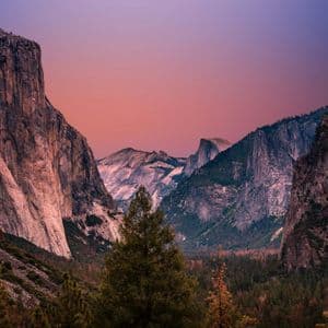 A vast mountain valley filled with a pine forest, flanked by steep granite cliffs under a purple and pink sky at dusk.