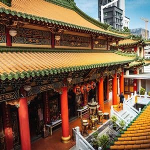 Un temple traditionnel orné avec des toits de tuiles orange et des piliers rouges, sur fond de bâtiments urbains modernes.