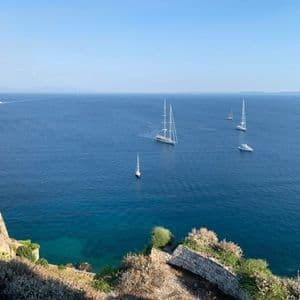 Blick von einer felsigen Klippe auf ein ruhiges, tiefblaues Meer mit mehreren Segelbooten und Yachten an einem klaren Tag.