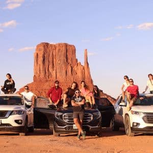Un grupo de viaje de WeRoad posa sobre y alrededor de tres SUVs en un desierto con imponentes formaciones de roca roja de fondo.