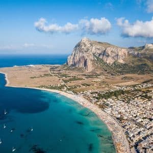 Vue aérienne d'une ville côtière avec une plage bondée le long d'une eau turquoise, à côté d'une grande montagne rocheuse sous un ciel bleu.