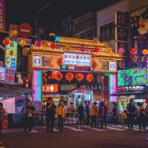 Des gens se tiennent à l'entrée d'un marché de nuit animé, encadré par une arche traditionnelle brillamment éclairée et des néons colorés.