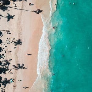 Una vista aerea dall'alto di una spiaggia sabbiosa accanto a un oceano turchese, con persone che nuotano e si rilassano sotto le palme.