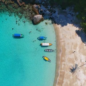 Vista aerea di barche ormeggiate in acque turchesi, con persone che nuotano vicino a una spiaggia sabbiosa e scogli.