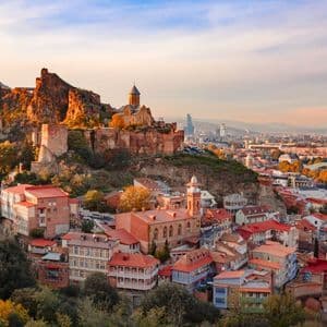 Una fortaleza histórica en una colina domina un casco antiguo con casas coloridas y el horizonte de una ciudad moderna al fondo.