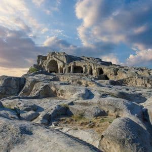 Una antigua ciudad troglodita excavada en una gran colina rocosa con un edificio de piedra en la cima, vista al atardecer.