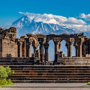 Las ruinas de un templo de piedra con columnas ornamentadas se alzan frente a una gran montaña nevada bajo un cielo azul parcialmente nublado.