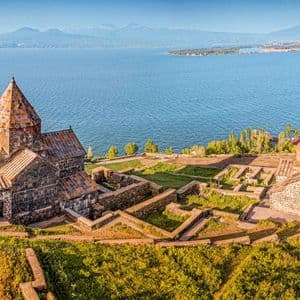 Un histórico complejo monástico de piedra en una colina verde, con vistas a un gran lago azul y montañas distantes en el horizonte bajo un cielo despejado.