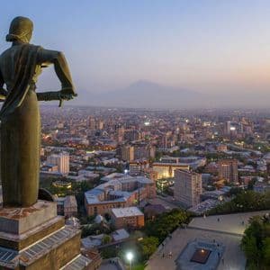 Una imponente estatua, vista de espaldas, se alza sobre una vasta ciudad iluminada al anochecer, con una montaña lejana en el horizonte.