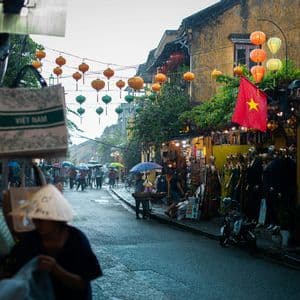 Des passants sous des parapluies déambulent dans une rue vietnamienne pluvieuse, bordée de boutiques et ornée de lanternes en papier colorées.