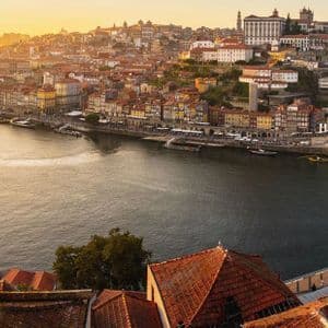Una vista panoramica di una città sul fiume con un grande ponte di metallo e edifici colorati illuminati dalla luce dorata del tramonto.