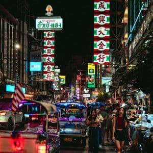 Una strada cittadina affollata di notte con persone che camminano e tuk-tuk nel traffico, illuminata da insegne al neon colorate con caratteri asiatici.