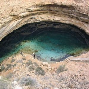 Un'ampia veduta di un grande cenote circolare pieno d'acqua turchese, con due persone in piedi sul bordo roccioso.
