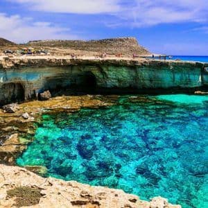 Scogliere rocciose con grotte marine incontrano un mare limpido e turchese, con persone in piedi sulla cima sotto un cielo azzurro.