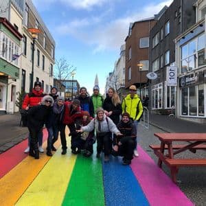 Un gruppo WeRoad in posa su una strada dipinta con i colori dell'arcobaleno, fiancheggiata da edifici e sotto un cielo parzialmente nuvoloso.