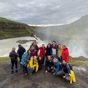 A WeRoad group trip posing for a photo on a clifftop next to a large waterfall and river canyon under a cloudy sky.