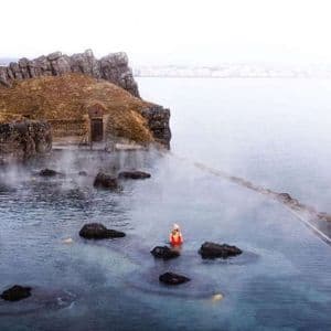 Une personne en maillot de bain rouge et bonnet se tient dans une piscine à débordement extérieure fumante, à côté d'une falaise rocheuse surplombant une vaste étendue d'eau.