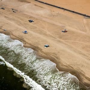 Une vue aérienne d'une plage de sable où l'océan rencontre le rivage, avec des véhicules et des tentes sur le sable et une route parallèle à la côte.