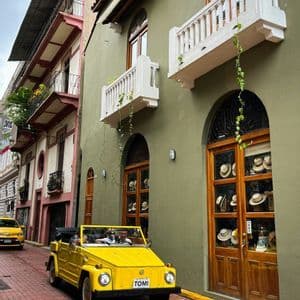 A yellow vintage convertible car drives on a red brick road next to colorful buildings with balconies.