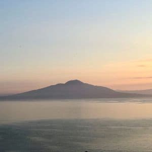 Ein großer Berg zeichnet sich vor einem pastellfarbenen Sonnenuntergangshimmel über einem ruhigen Gewässer ab.