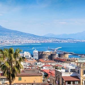 Vista dall'alto di una città costiera con un grande vulcano sullo sfondo, che mostra tetti, una palma e una baia blu brillante.